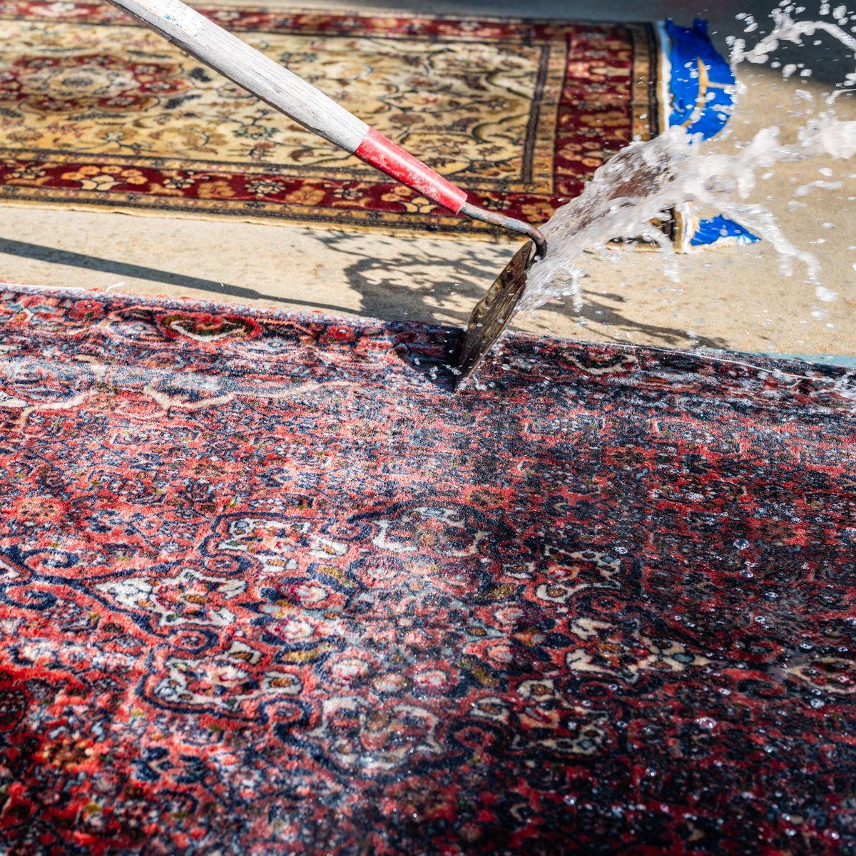 A water jet from a spray wand cleans a large, patterned rug on a concrete surface.
