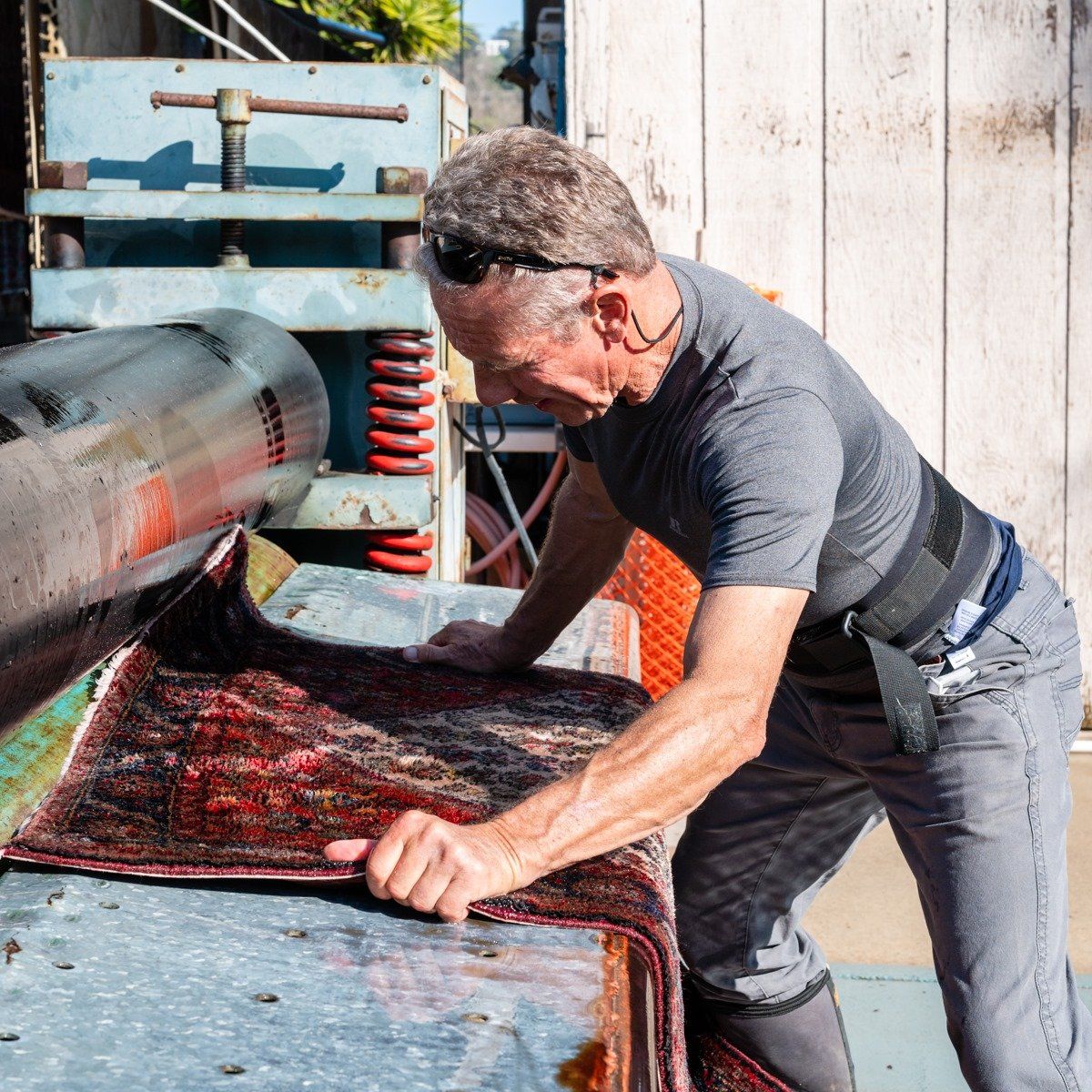 A person in a gray shirt uses a large industrial press to flatten a dark, patterned rug in a workshop.