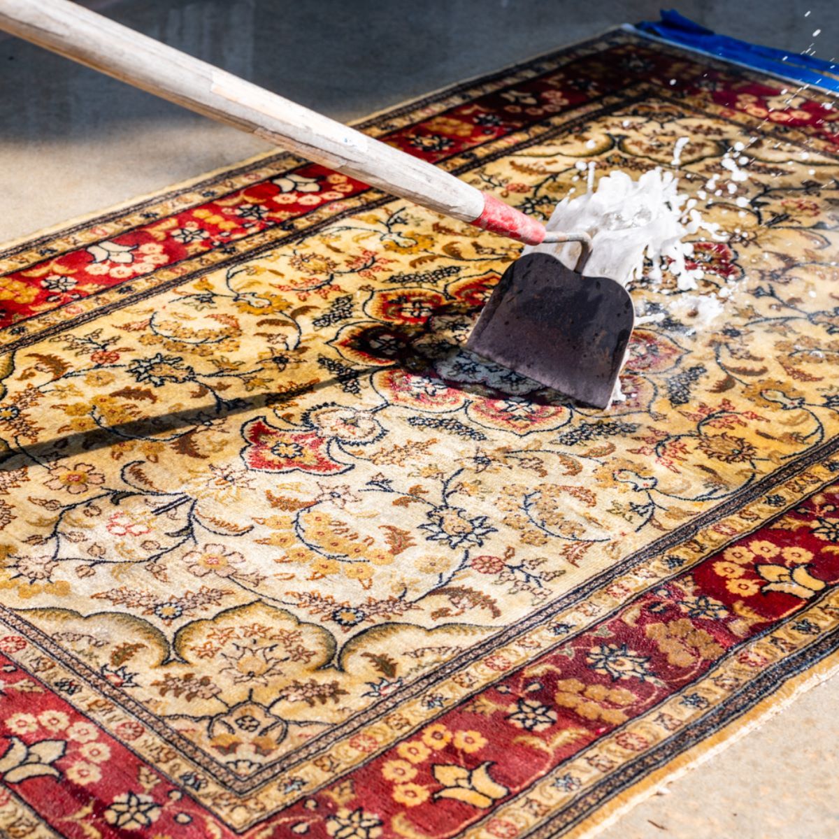 A hoe tool is being used to scrub white suds across a patterned, vintage-style area rug.