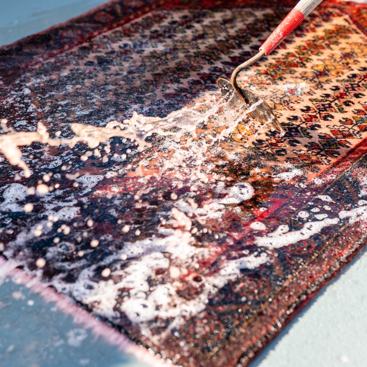 A water hose sprays a stream of soapy water onto a patterned Persian-style rug lying on a flat, light-colored surface.
