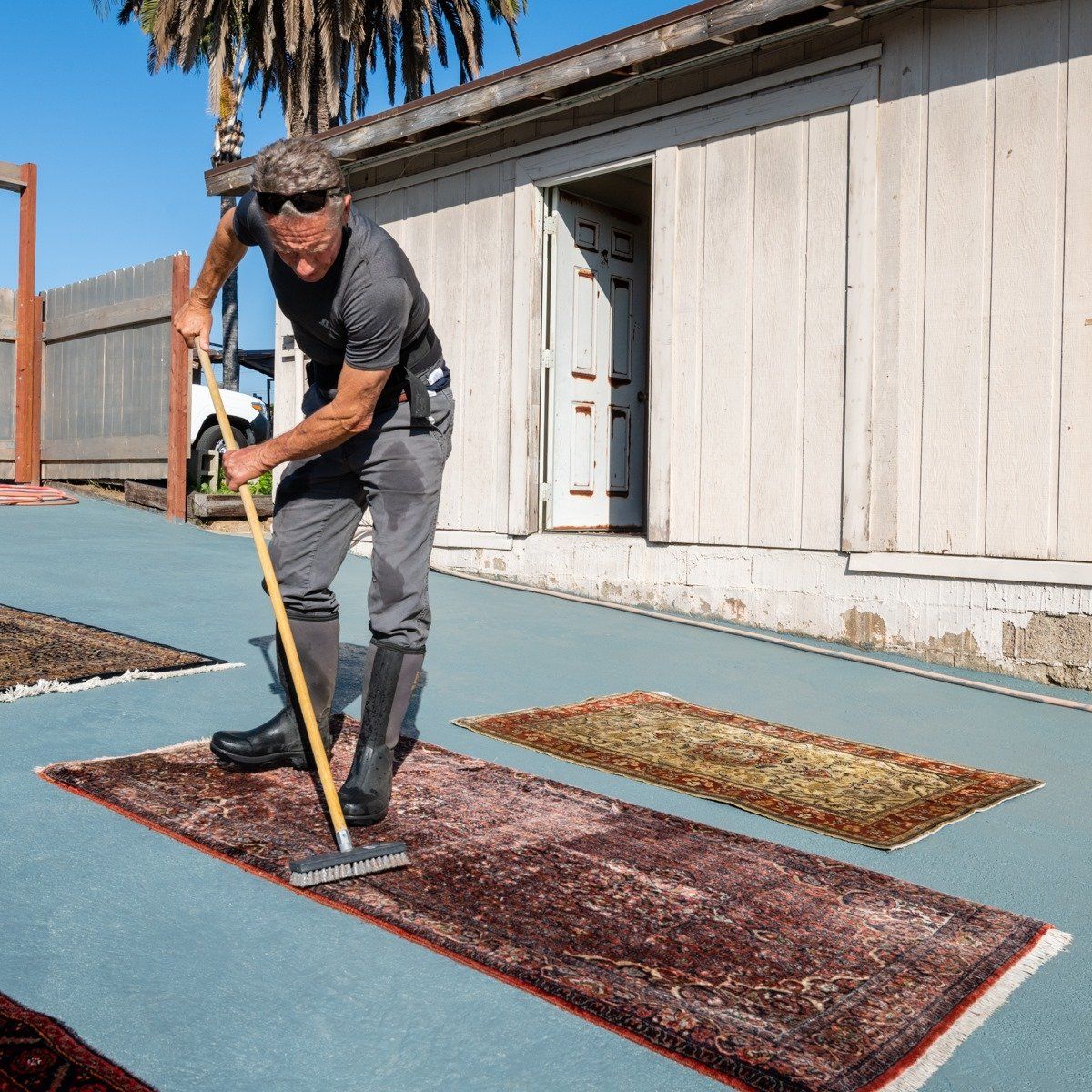 A person wearing sunglasses and boots uses a long-handled brush to clean a rug on an outdoor blue surface.