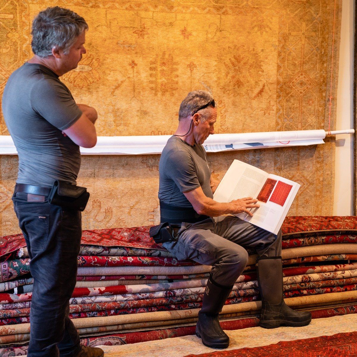 Two people in grey shirts and dark trousers stand and sit in a room filled with stacked rugs, reviewing a book.