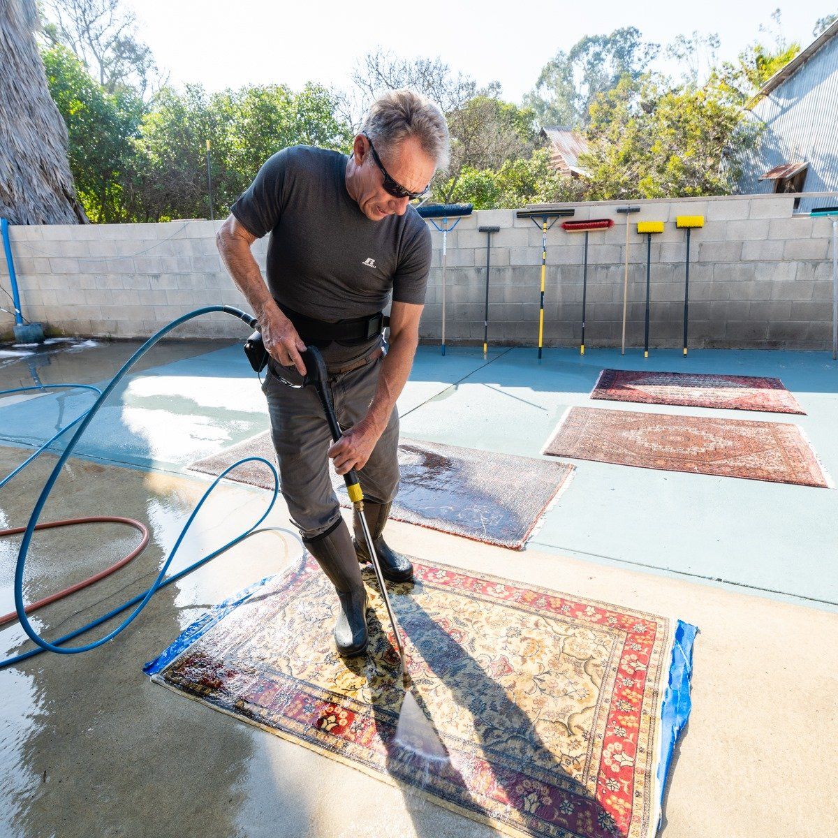 A person in a gray shirt and boots cleans an oriental rug on a concrete patio using a water hose tool.