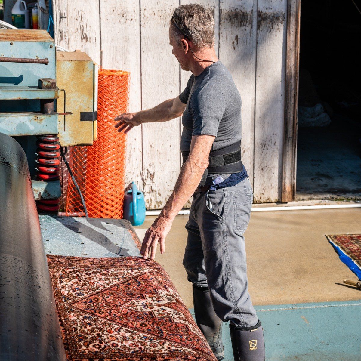 A person wearing a back brace and boots stands in an industrial space, feeding a patterned rug into a large machine.