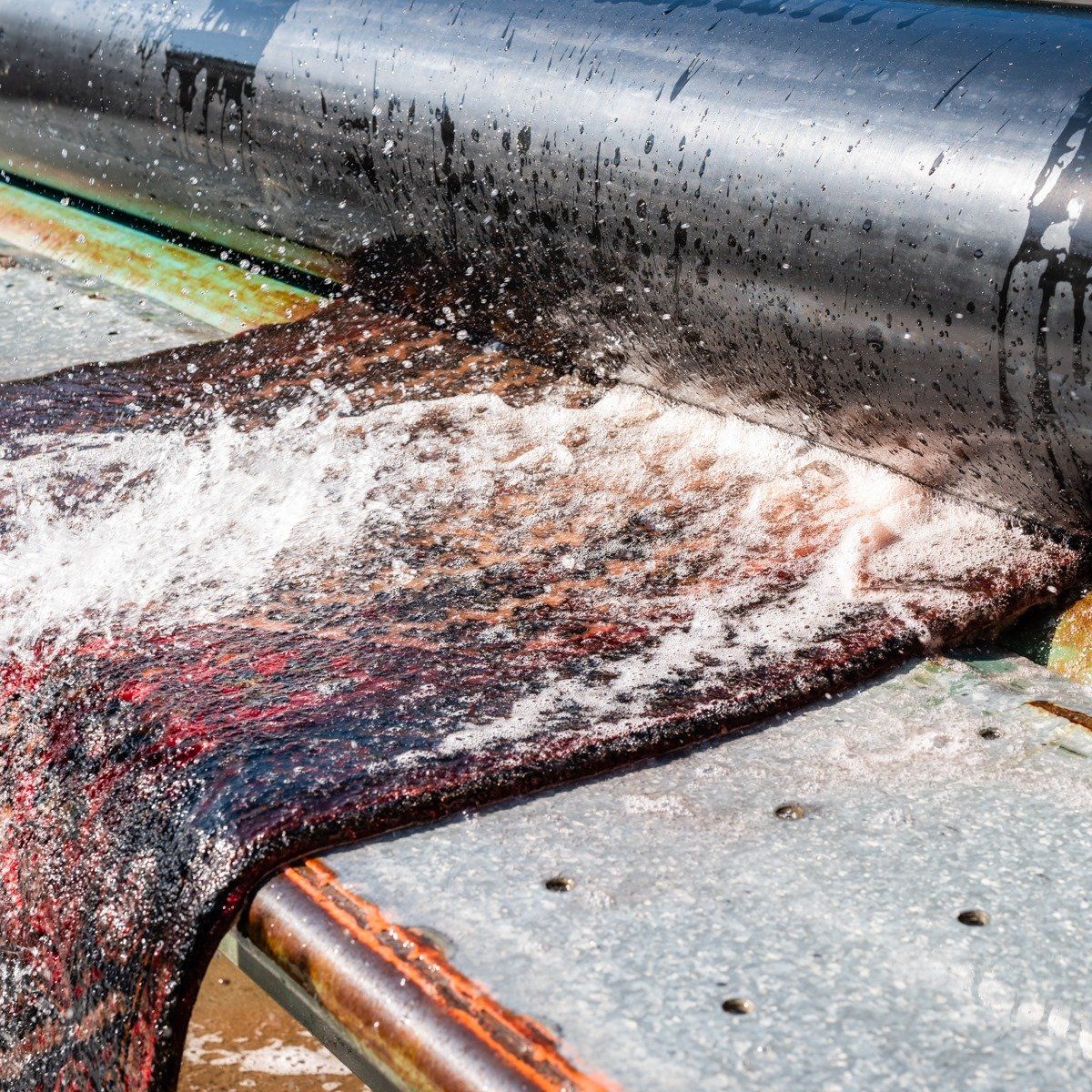 A dark, textured material is being processed under a large, wet metal roller at an industrial facility.