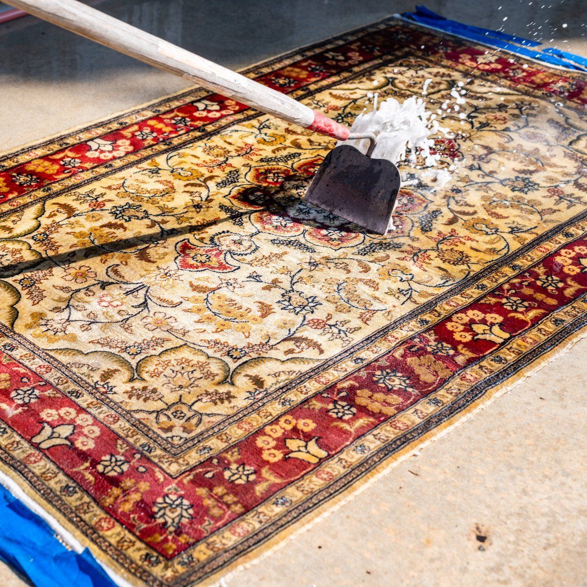A garden hoe is being used to scrub soap suds onto an ornate, cream and red patterned rug laid on a concrete surface.