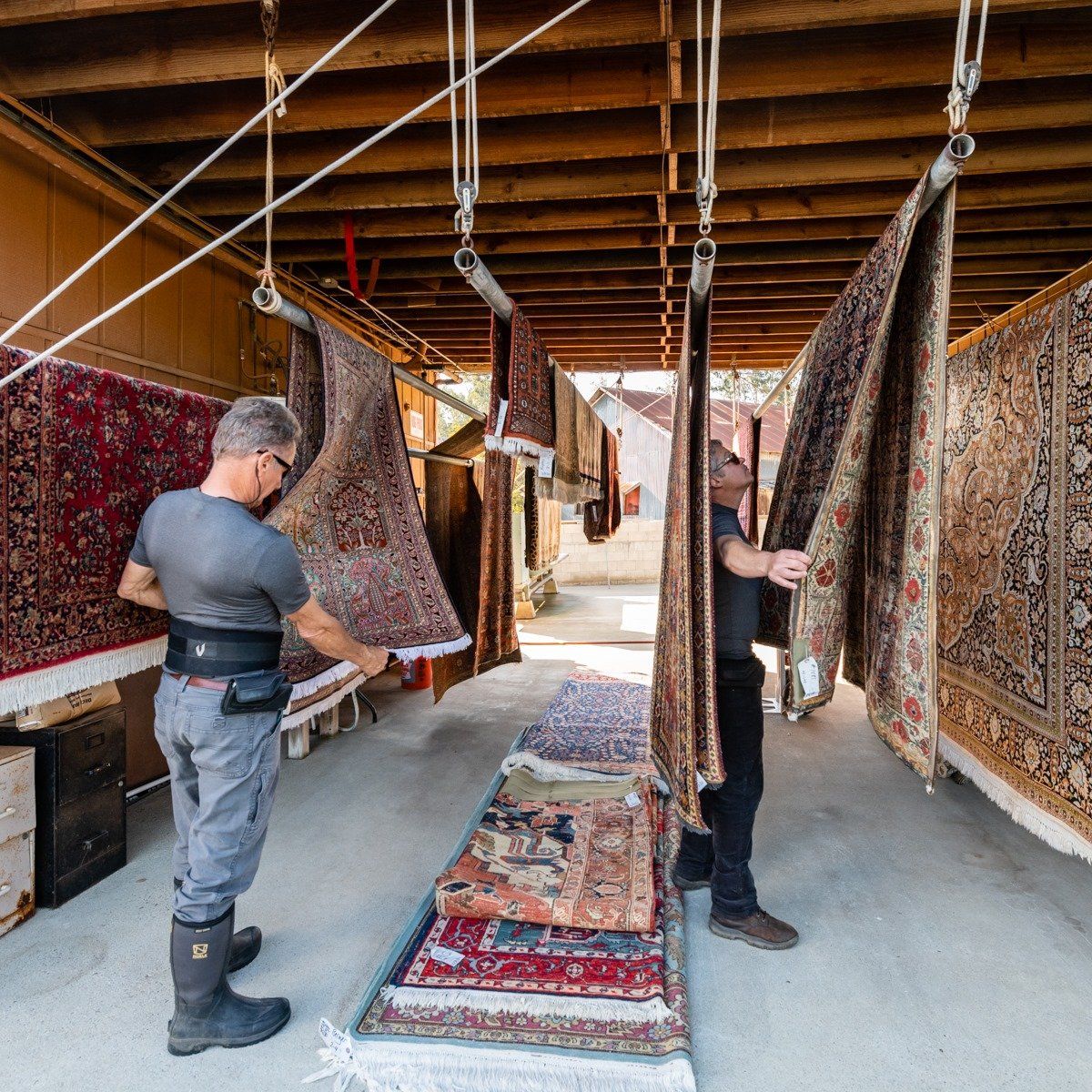 Two workers hang several large, intricate, patterned area rugs to dry in a covered outdoor facility.