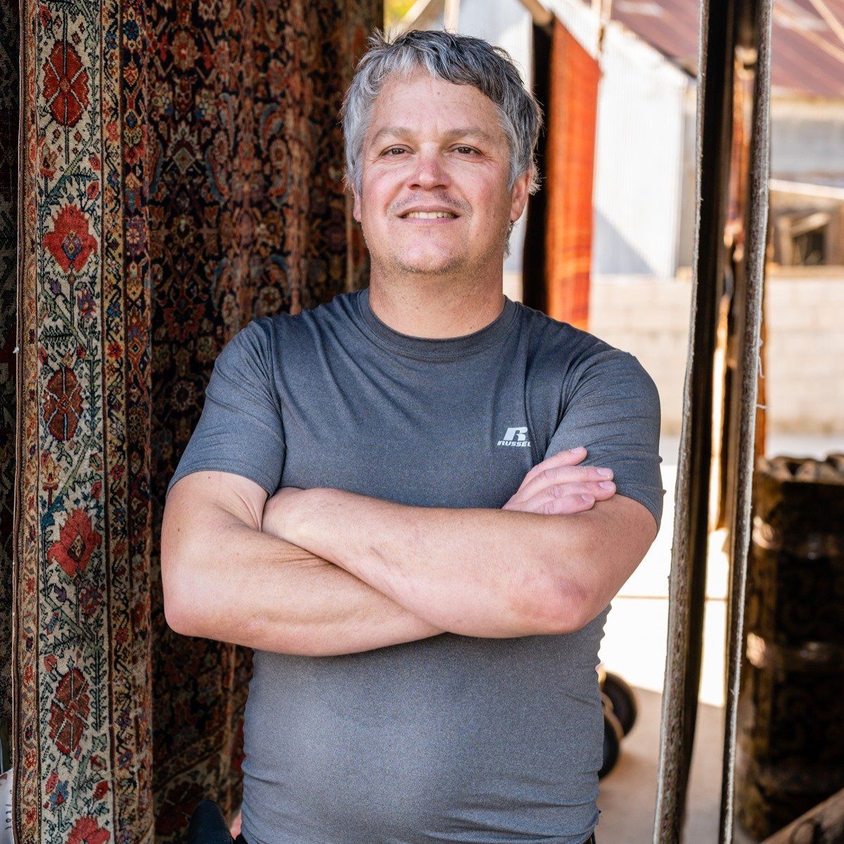 A smiling person in a grey t-shirt stands with arms crossed, positioned in front of a patterned, hanging textile.
