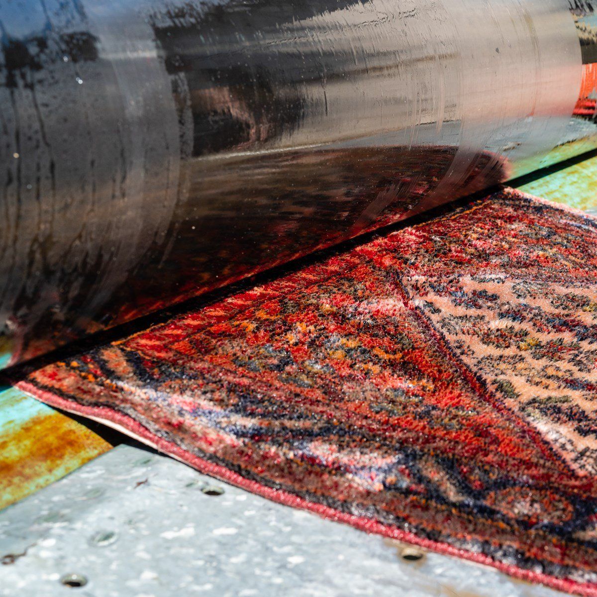 A patterned rug with red and black tones passes through a large industrial roller in a manufacturing setting.