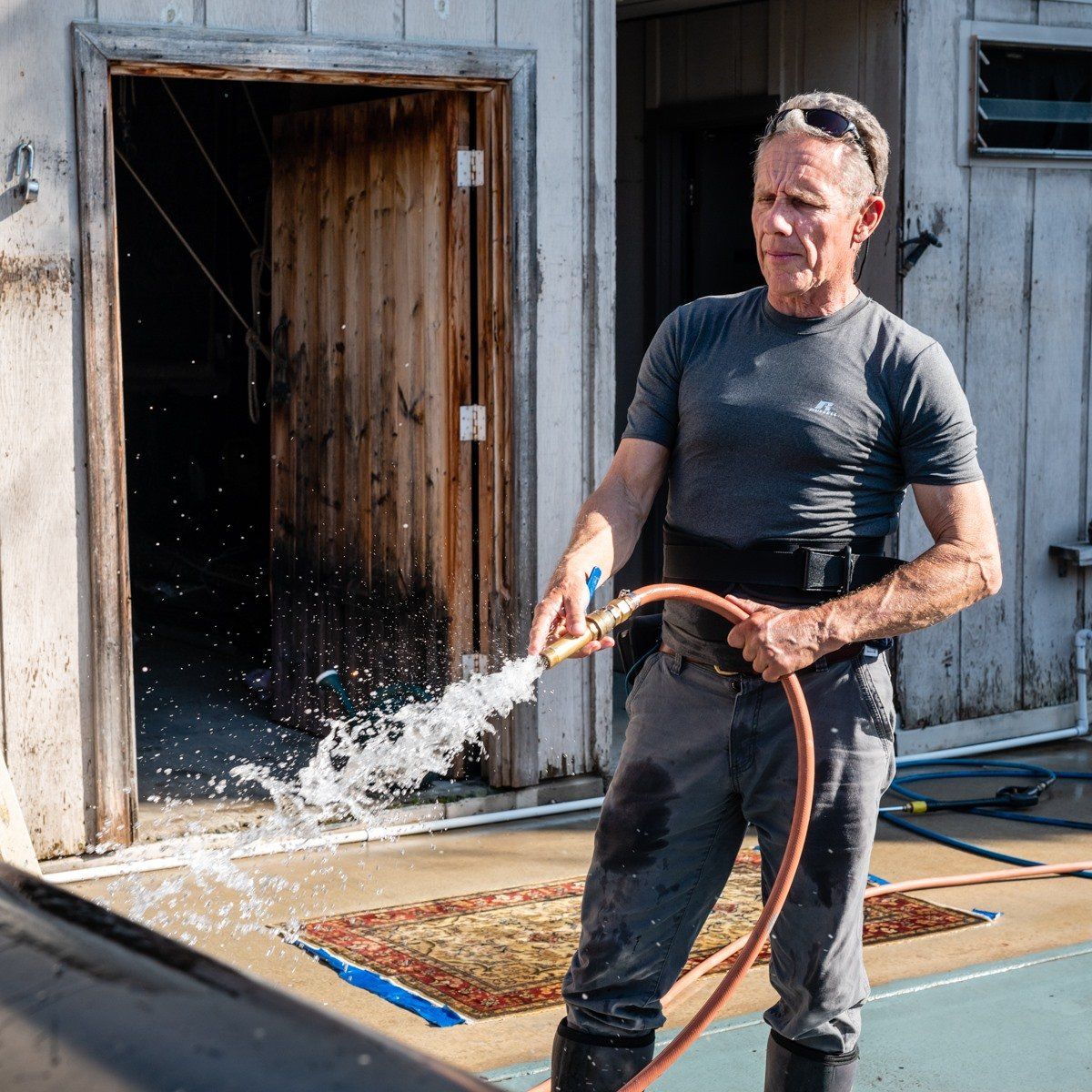 A person in a gray t-shirt and work pants sprays a water hose in front of an open doorway and a patterned rug.