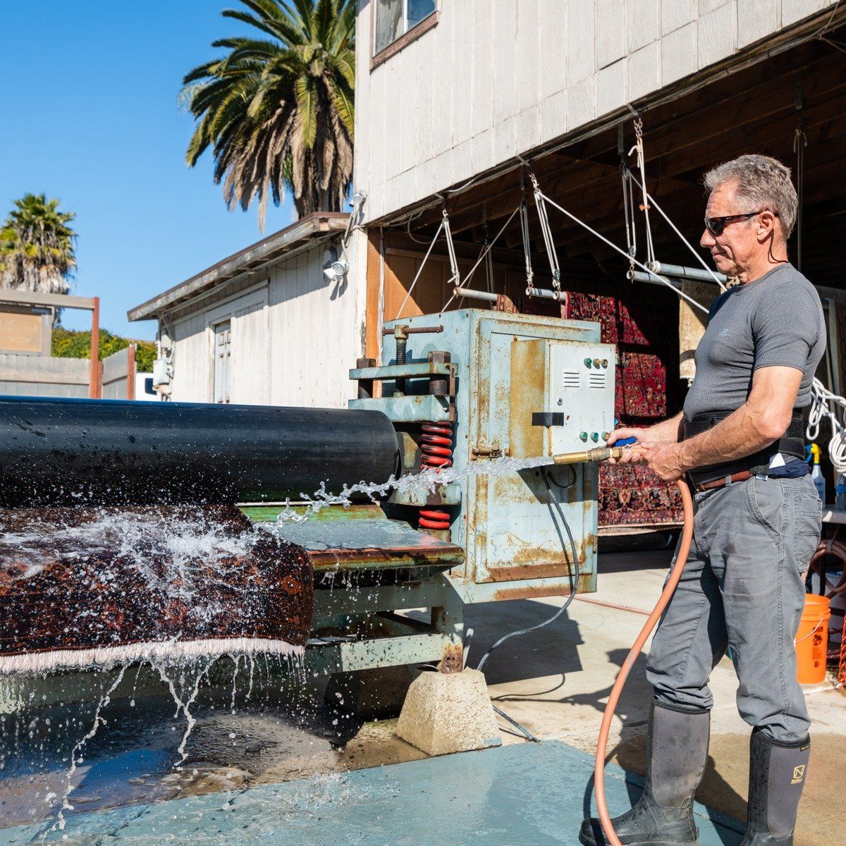 A person in a gray shirt and boots uses a hose to spray water onto a dark, flat material being processed by a machine.