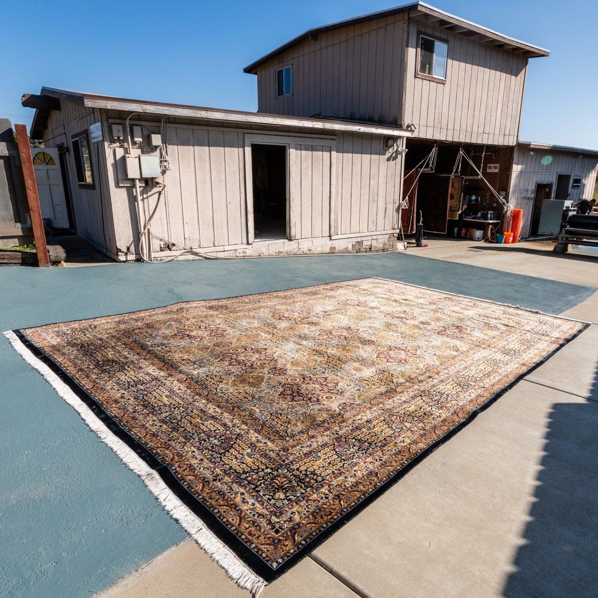 A large, rectangular, patterned area rug in shades of tan, brown, and blue displayed outdoors on a paved area.