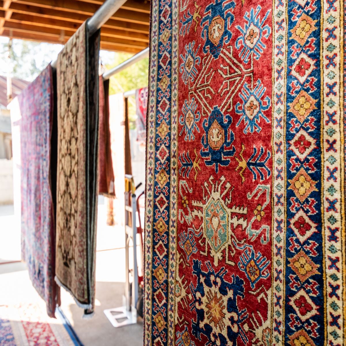 Several colorful, intricately patterned woven rugs hang from a display rack in an outdoor setting.
