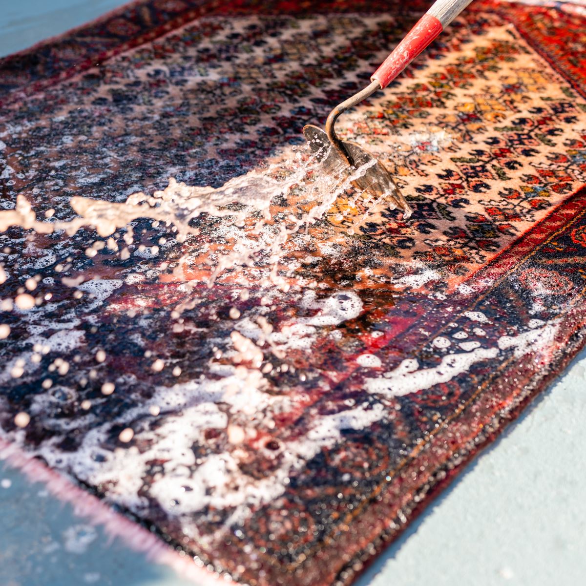 A rug being cleaned with soap and a brush on a flat surface, with white suds visible on its dark patterned fabric.