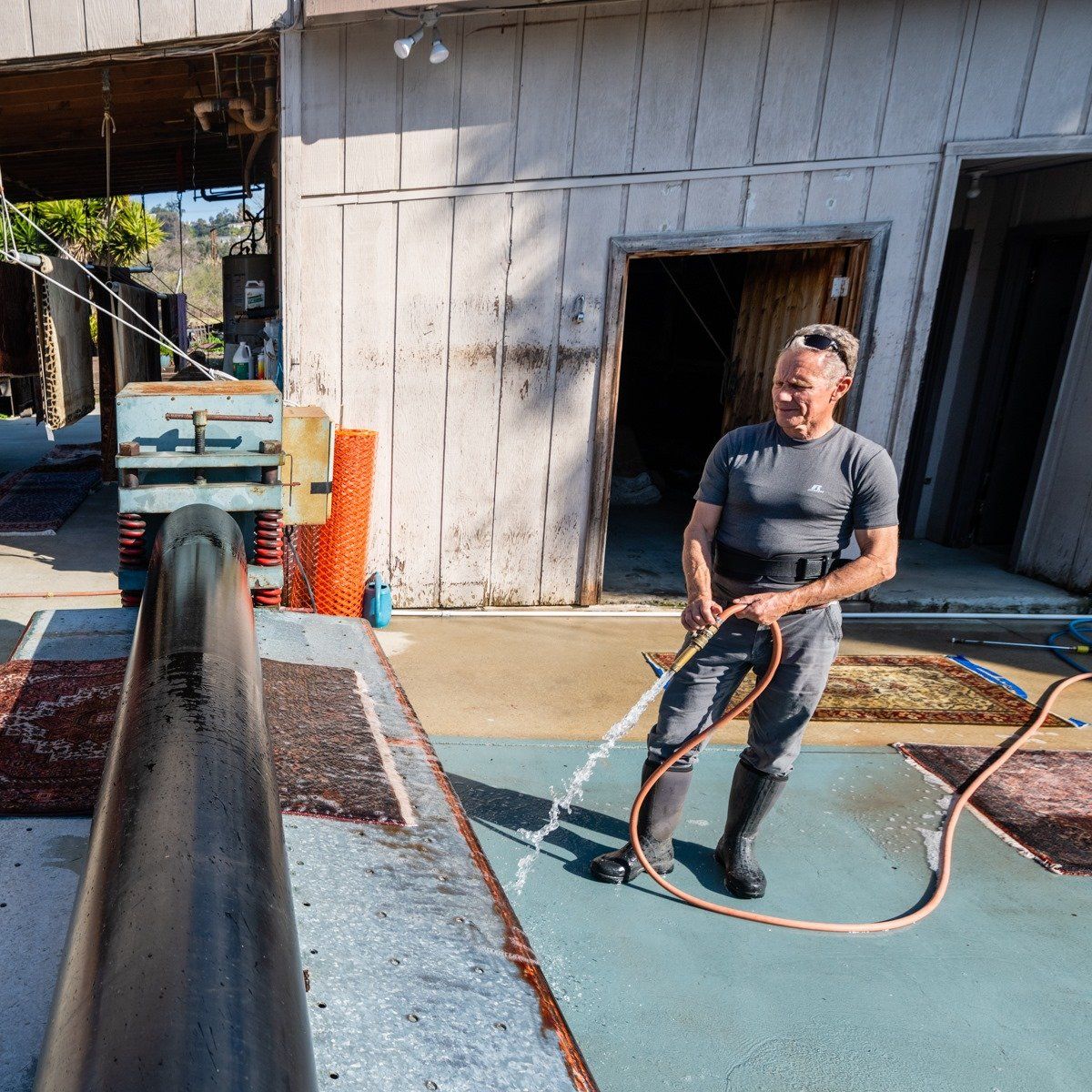 A person in a gray shirt and dark pants stands outside, spraying water from a hose toward a long, black metal pipe.