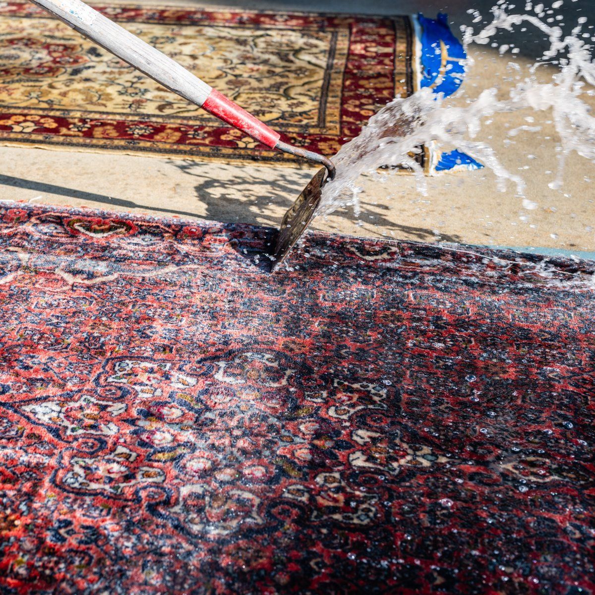 A pressure washer sprays water onto a red patterned area rug on a concrete surface, with a second rug visible nearby.