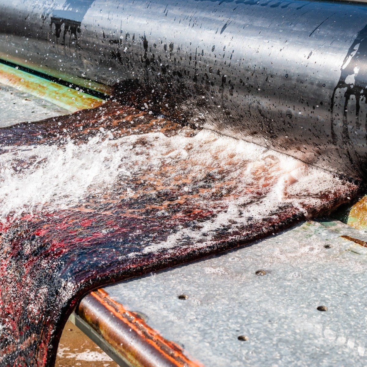 Water flows over a dark, textured textile material as it passes under a smooth, cylindrical industrial roller.