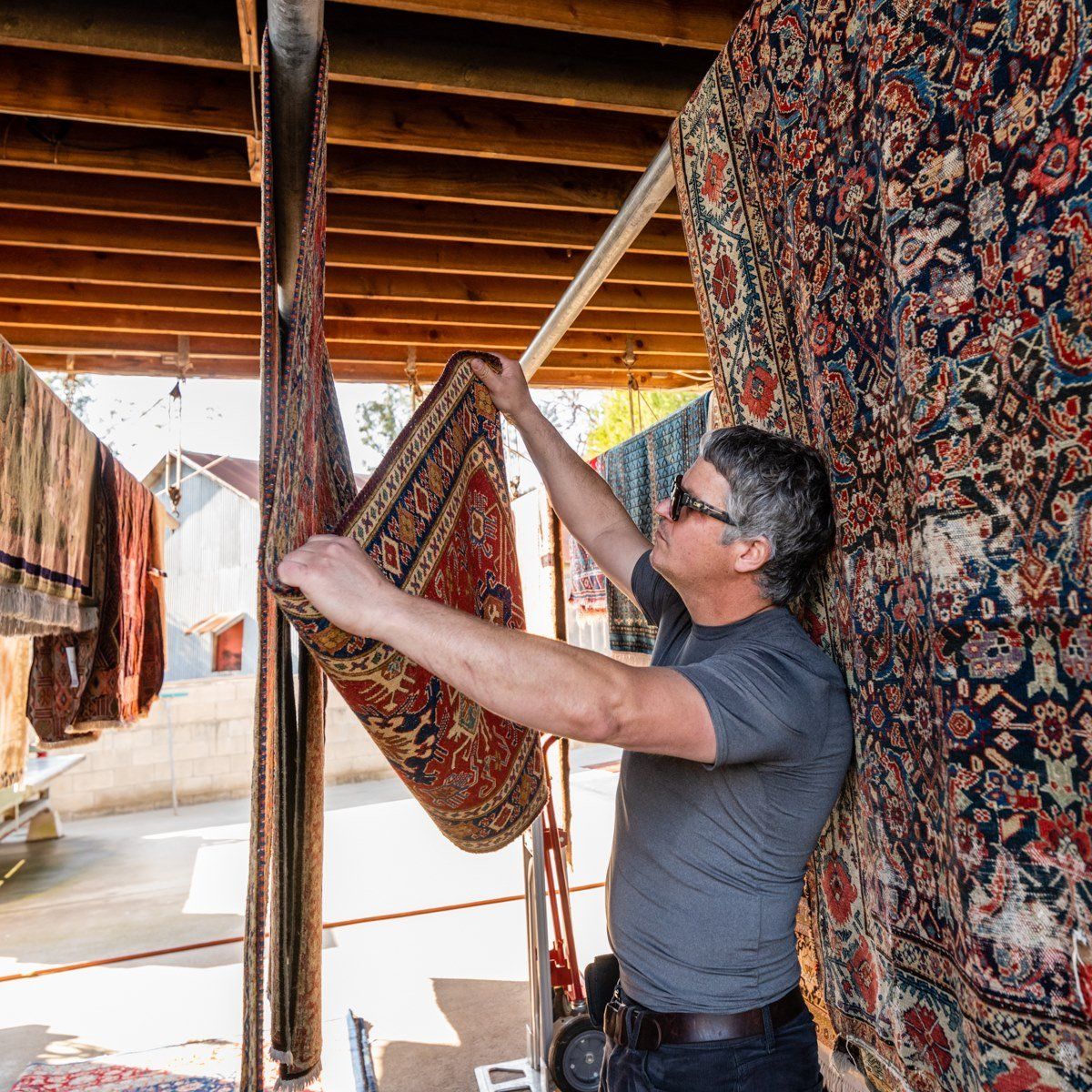 A person in sunglasses examines a red patterned rug hanging from a rack in an outdoor display area.