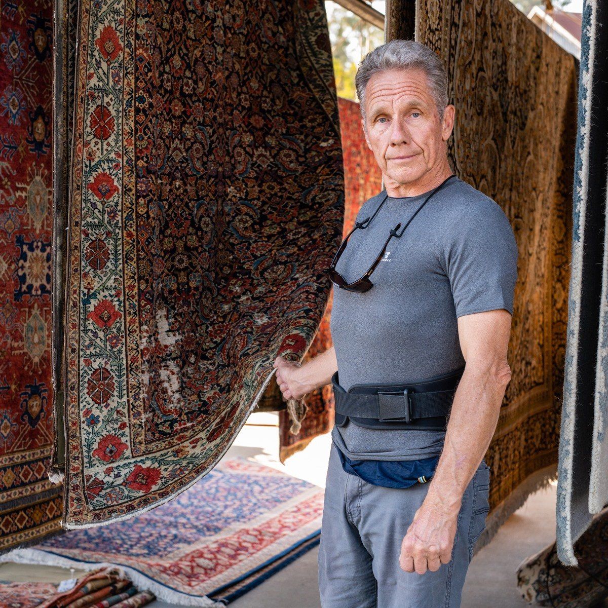 A person stands in a warehouse filled with hanging decorative rugs, holding the edge of one dark patterned rug.