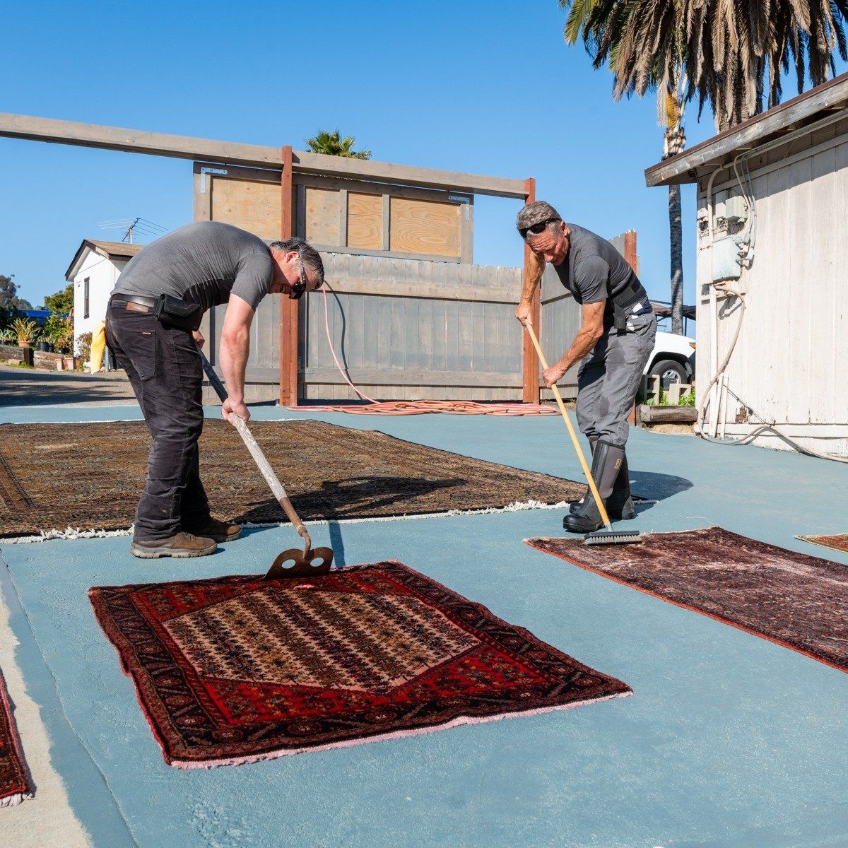 Two people use long-handled tools to clean several patterned rugs laid out on a flat, blue outdoor surface.