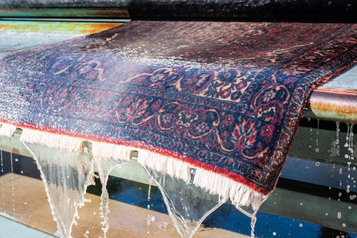 A dark, patterned rug with white fringe is being thoroughly rinsed with water in a professional cleaning facility.