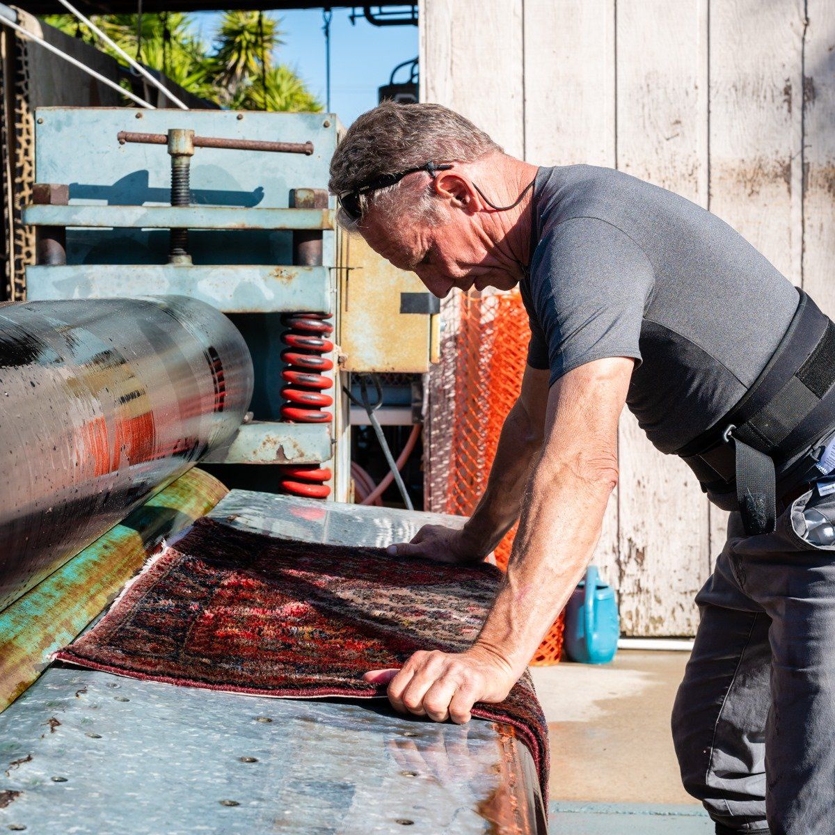A person wears a back brace while feeding a patterned rug into a heavy-duty industrial wringing machine.