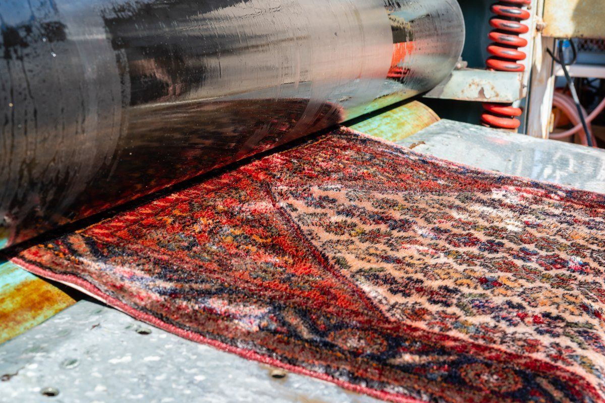 A decorative, patterned rug passes beneath a large metal industrial roller during a manufacturing or cleaning process.