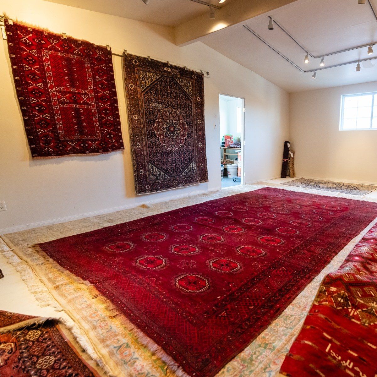 A gallery space featuring two large rugs hanging on a white wall and a large red patterned rug spread on the floor.