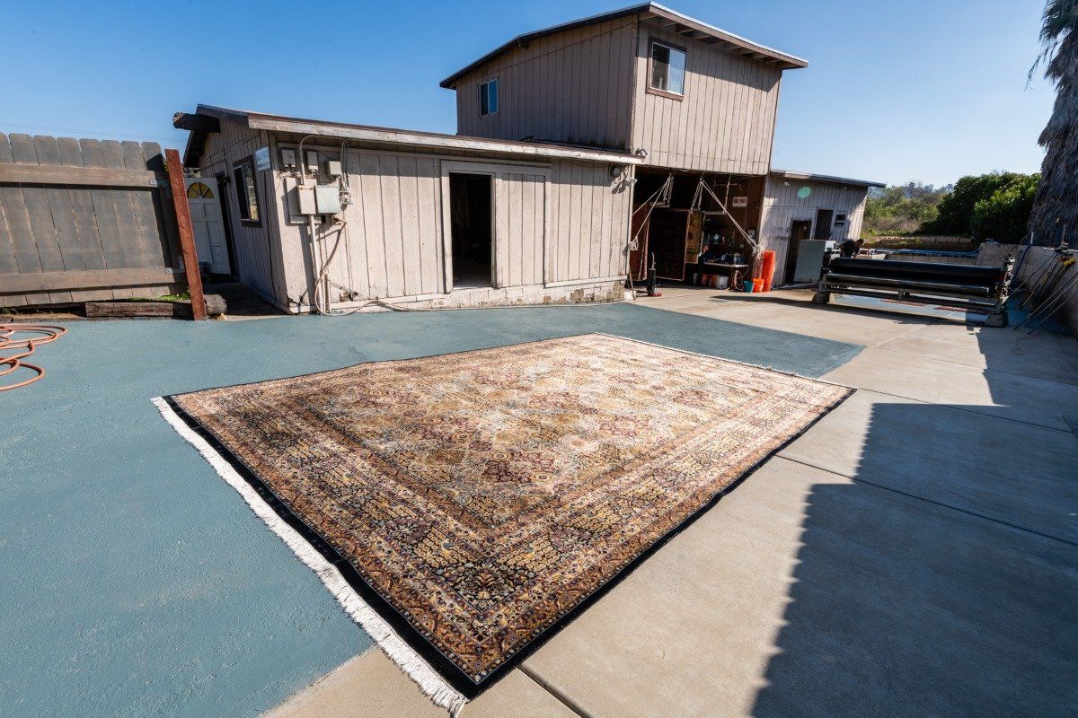 A large, intricately patterned, neutral-toned rug laid out on a blue concrete patio in front of a rustic wooden building.