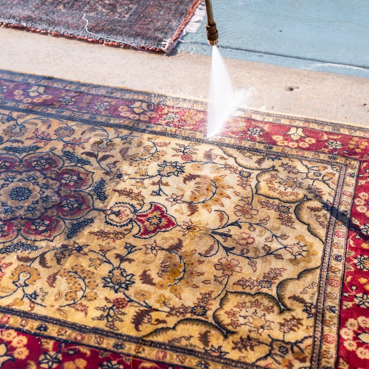 A high-pressure water nozzle sprays and cleans an intricate, patterned Persian-style rug on a concrete surface.