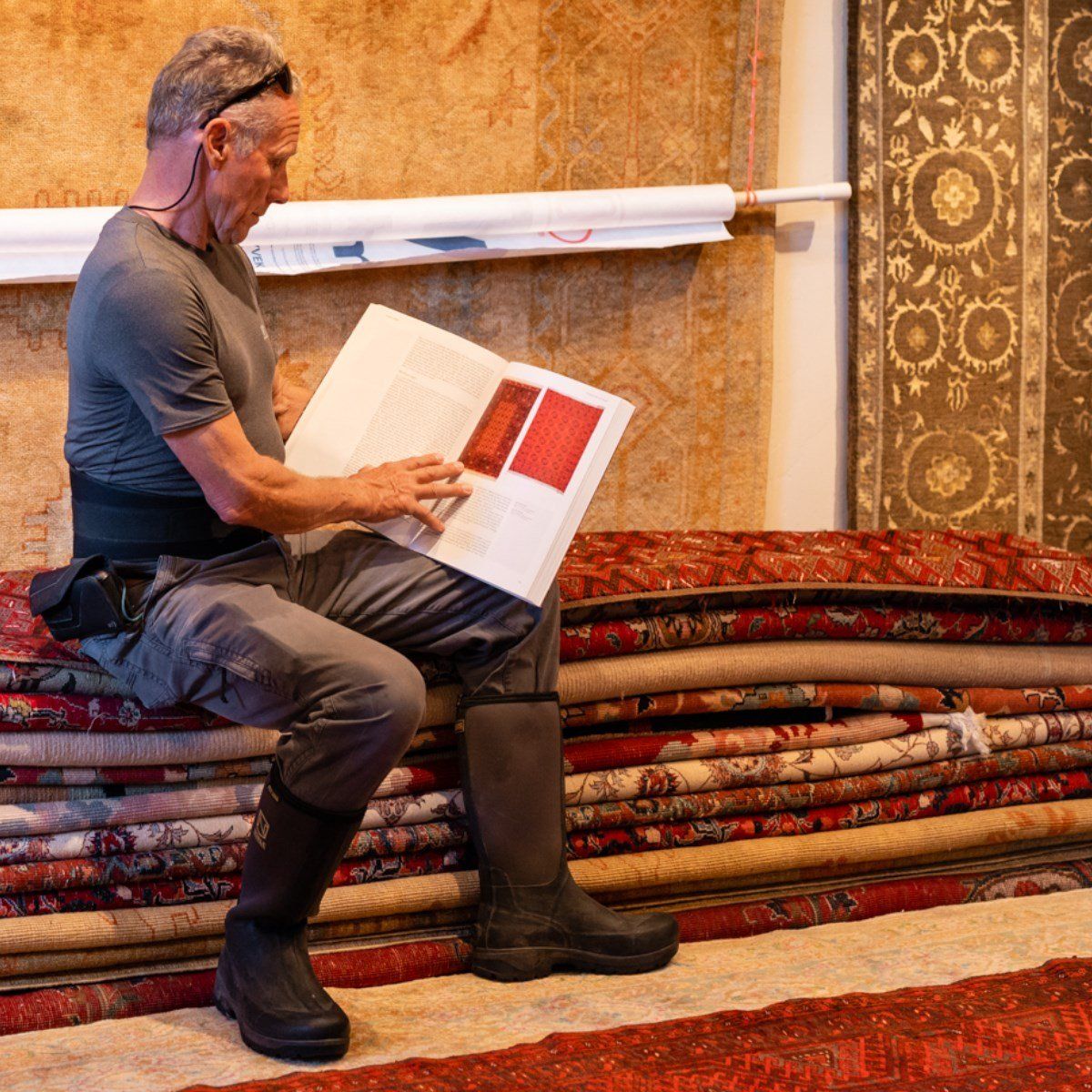 A person in a gray shirt and boots sits on a stack of rolled rugs, reading an open book in a room filled with carpets.