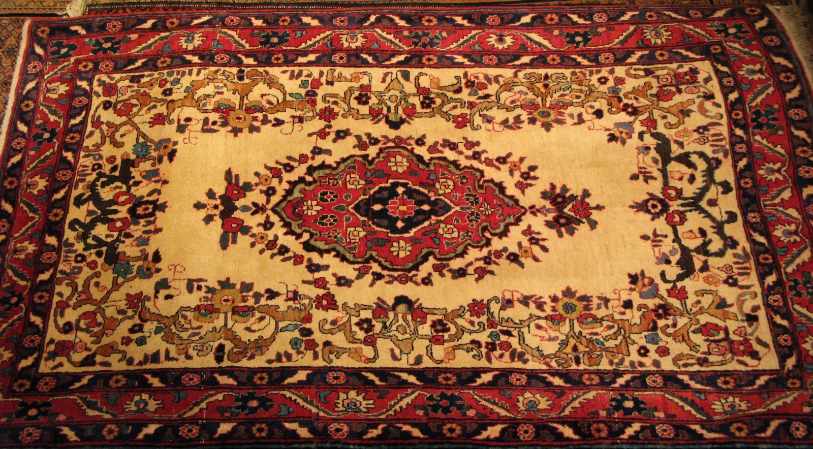 An ornate, rectangular Persian-style rug with a cream center, a central floral medallion, and a patterned red border.