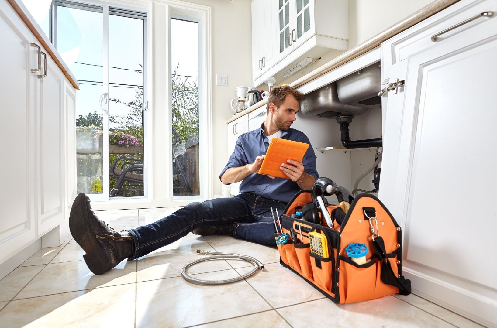 A man is sitting on the floor in a kitchen looking at a tablet.