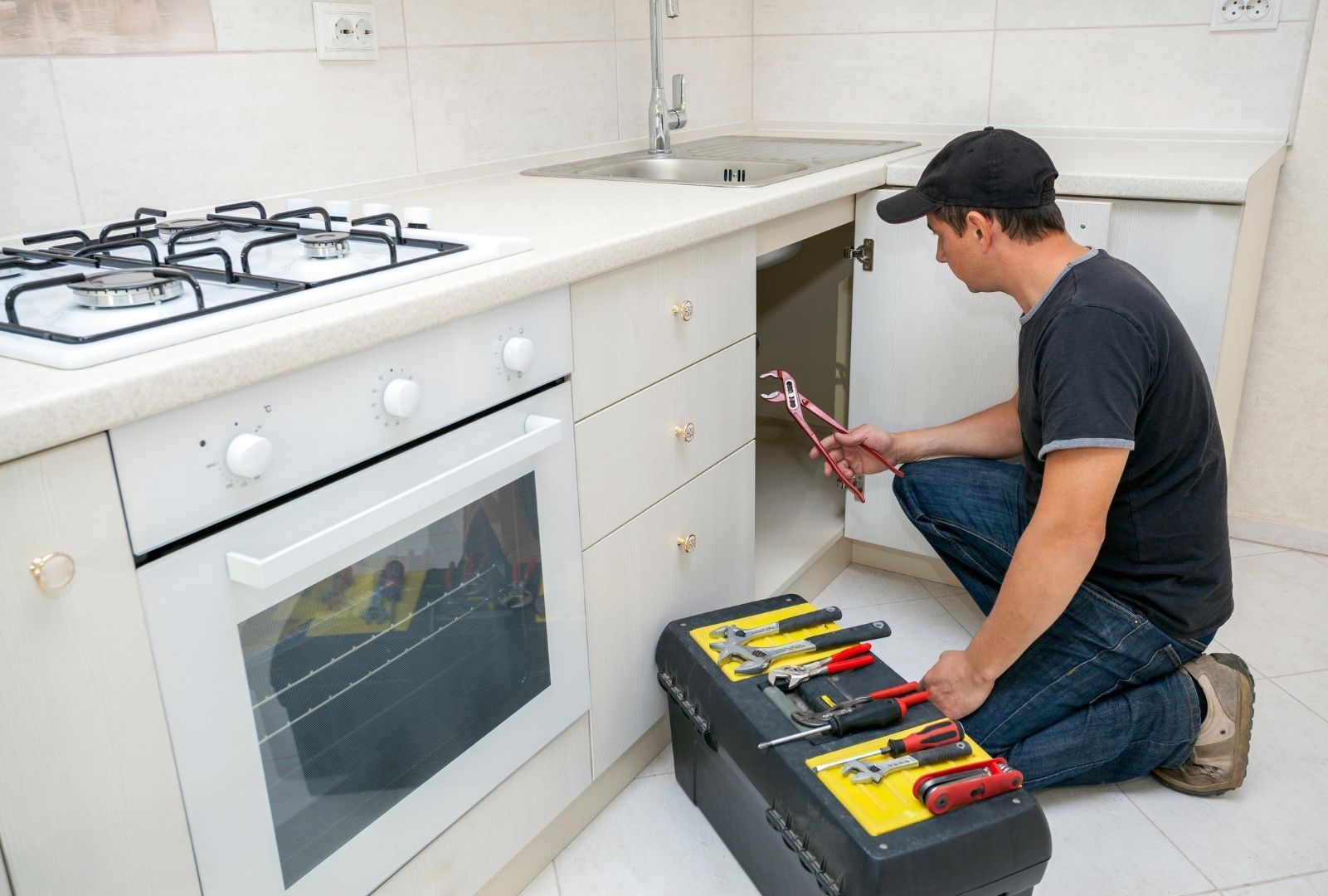 A man is kneeling down in a kitchen with a toolbox and a wrench.