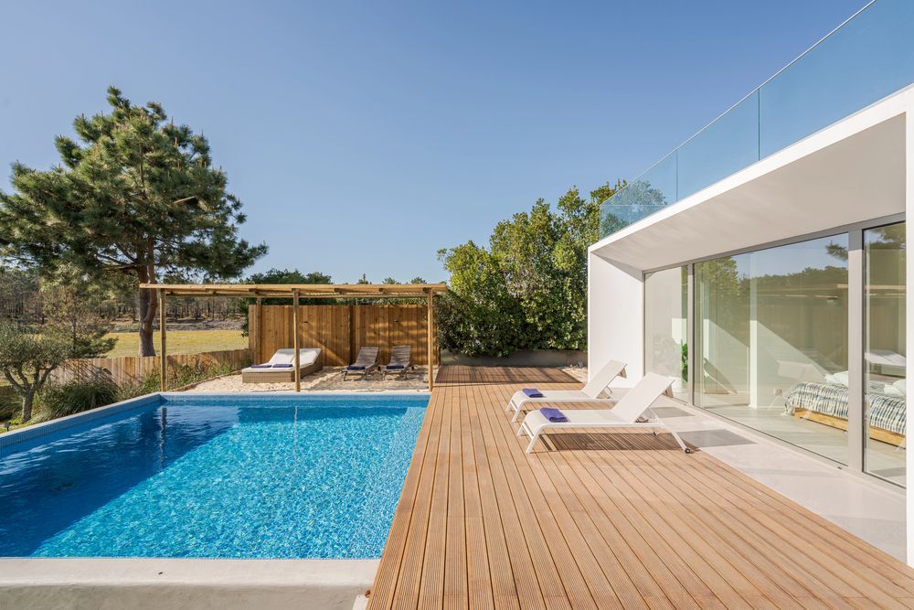 Scena a bordo piscina: piscina blu, terrazza in legno, sedie a sdraio, moderno edificio bianco, giornata di sole.