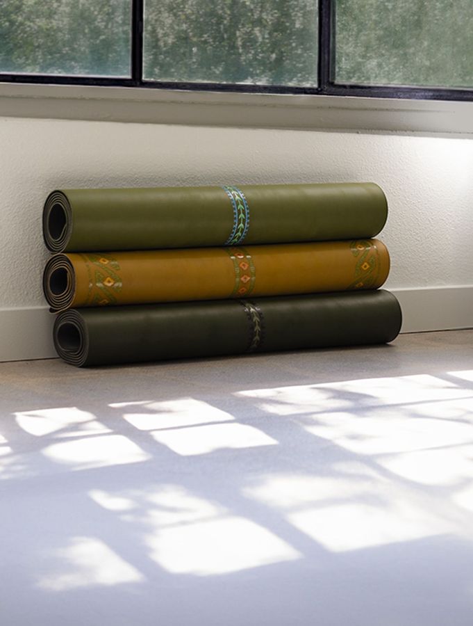 Three rolled yoga mats in olive, gold and forest green leaning against the wall beneath a window at Yoga Shala Collective, with morning light and window-pane shadows across the concrete floor