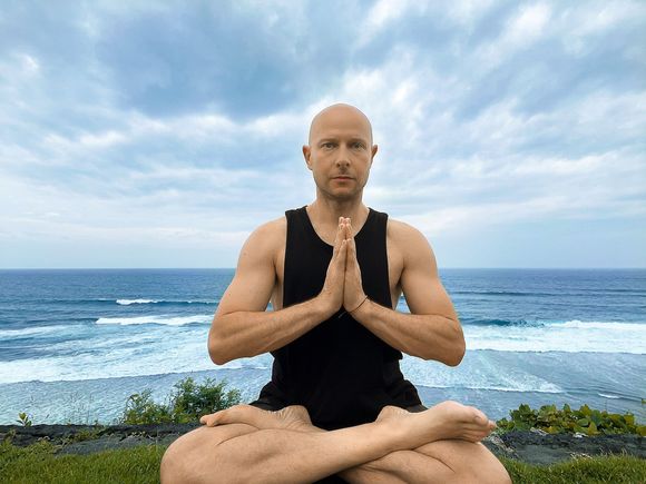 David Royer, founder of Yoga Shala Collective, seated in lotus pose with hands at heart centre on a clifftop above the ocean