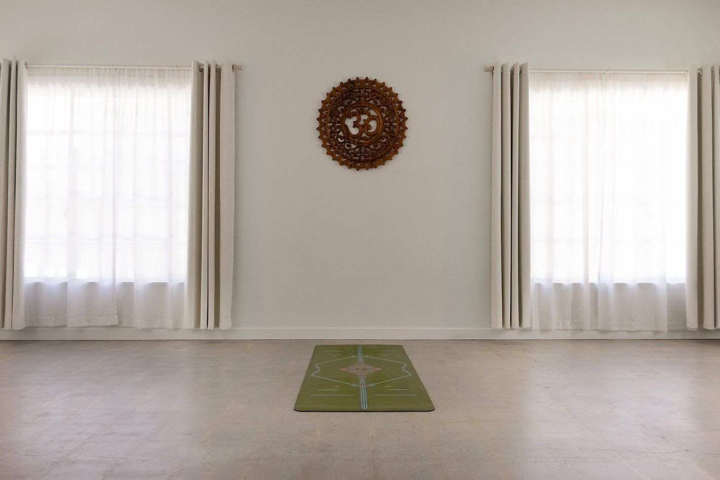 Practice room at Yoga Shala Collective with a carved wooden om medallion centred on the wall, two curtained windows and a single green yoga mat on the concrete floor