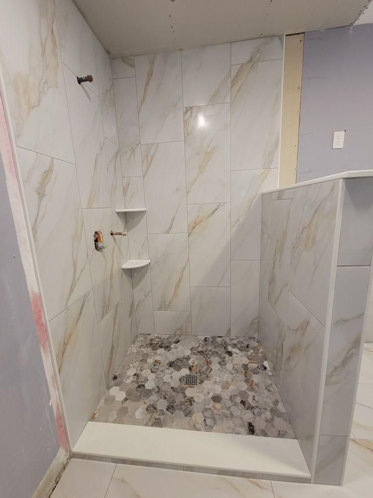 Tiled walk-in shower with marble-look walls, built-in corner shelves, and pebble stone floor.