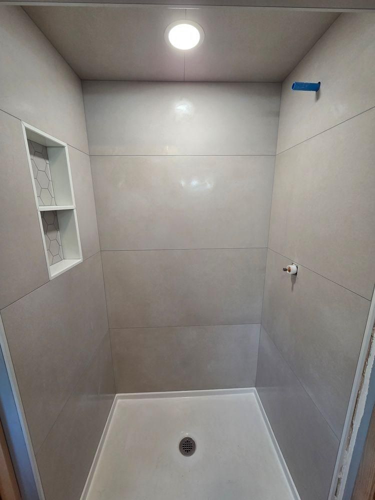 Small tiled shower stall with a built-in shelf, drain, and overhead light