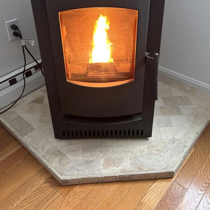 Black pellet stove with visible flame on a stone hearth beside a hardwood floor and wall outlet