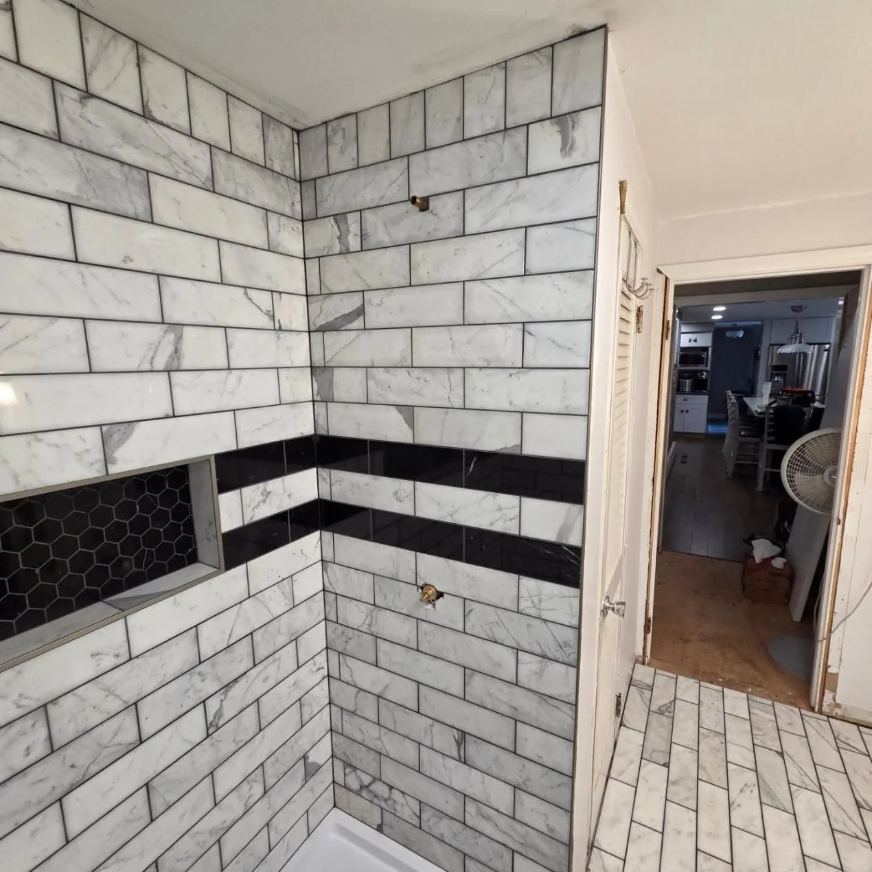 Tiled shower stall with white subway tiles, black accent band, and doorway to a bathroom or utility area