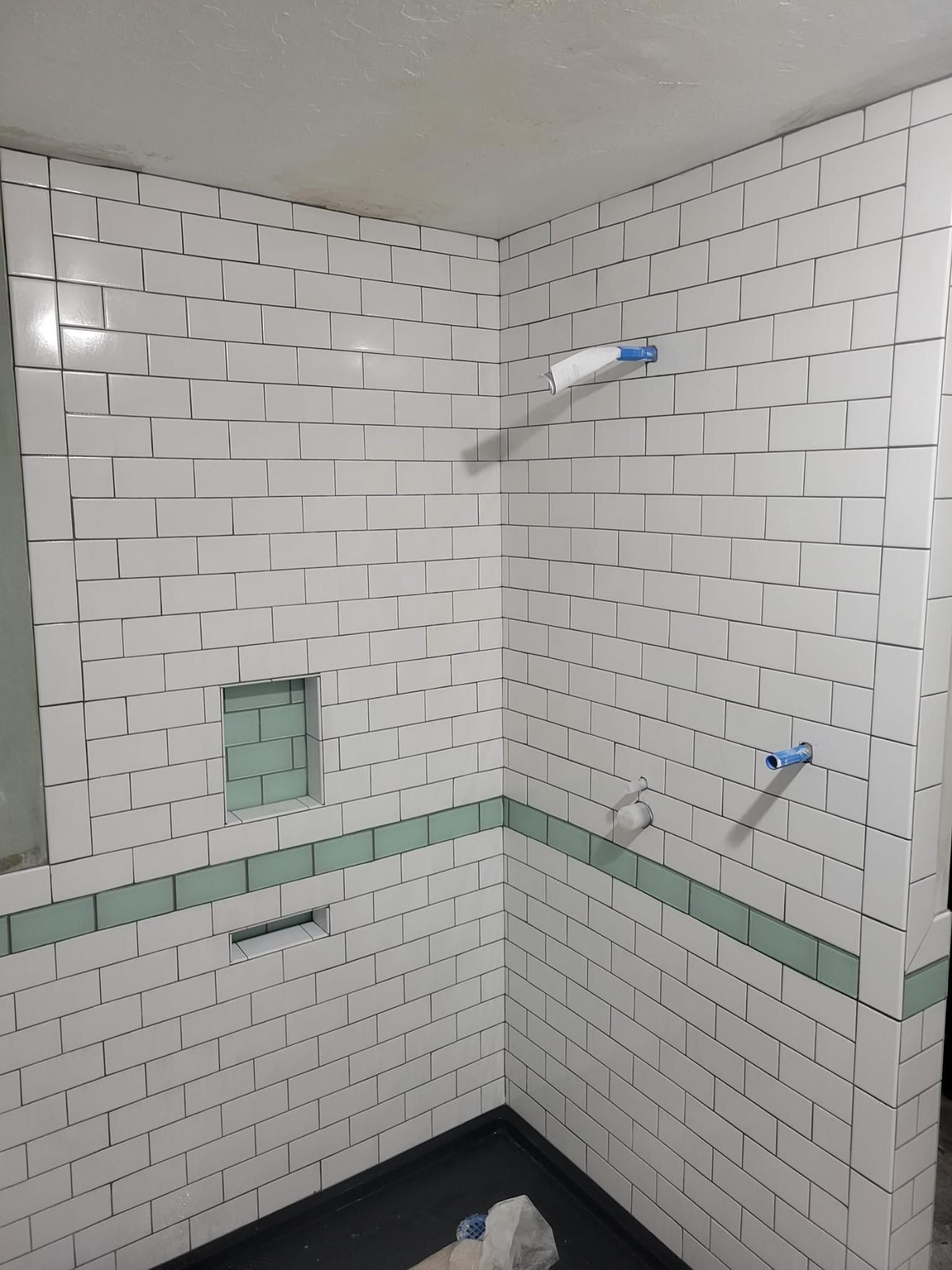 Tiled bathroom shower corner with white subway tiles, green accent band, and two blue pipe fittings in the wall