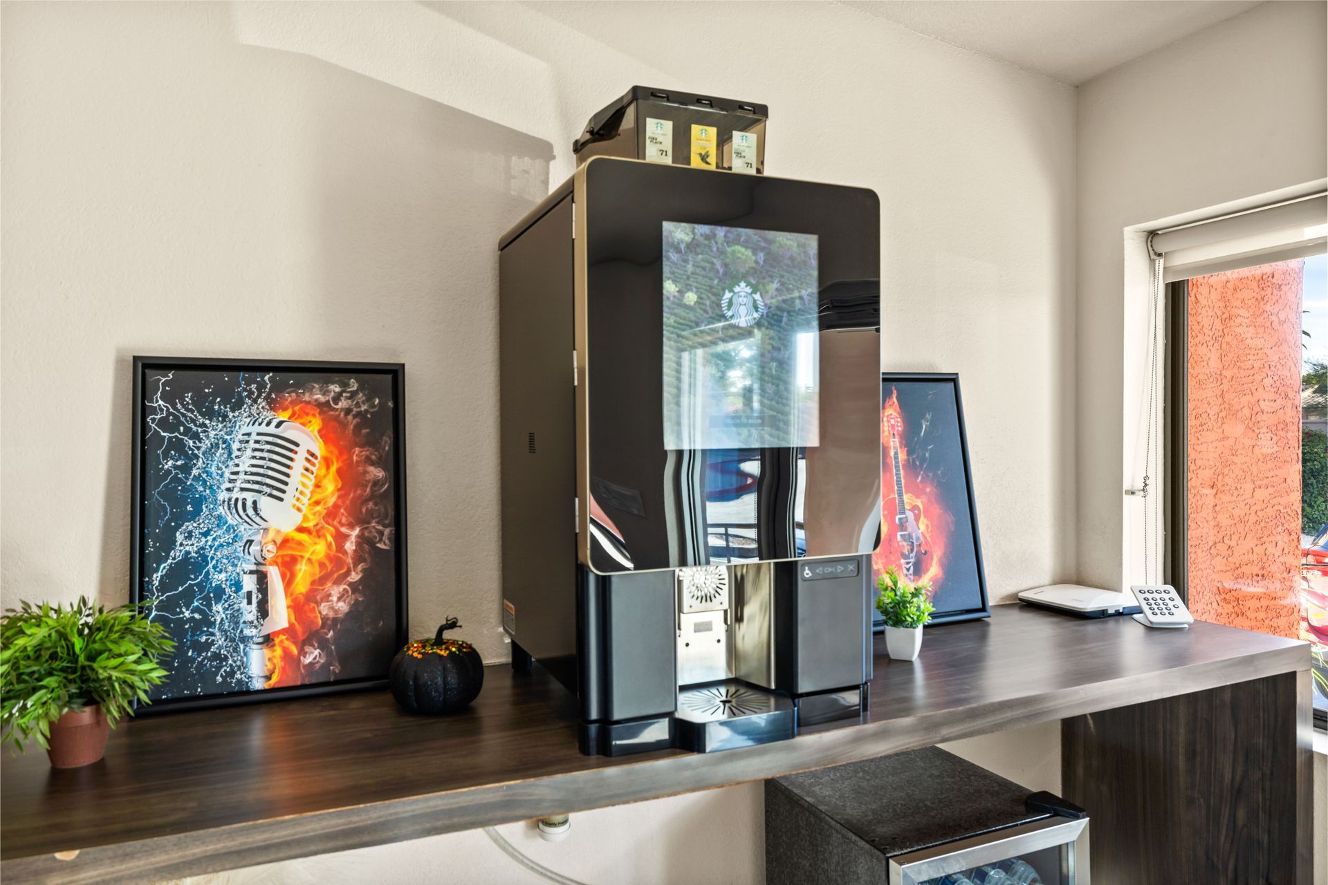 A large black coffee machine sits on a wooden desk with artwork, plants, and a window in the background.