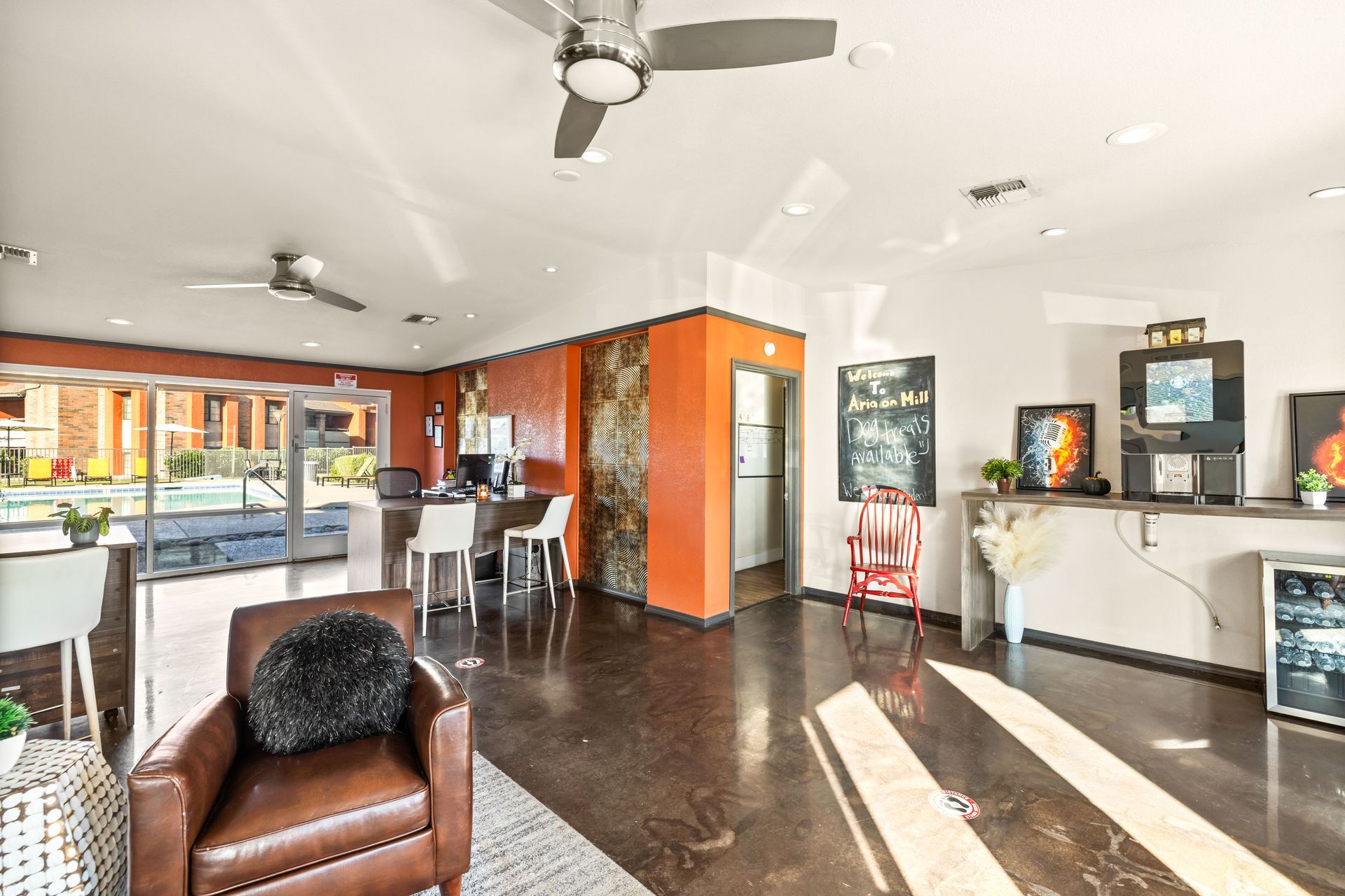 Lobby with seating, desk, and refreshments. Orange and white walls, dark floor, ceiling fans, and view to an outdoor pool.