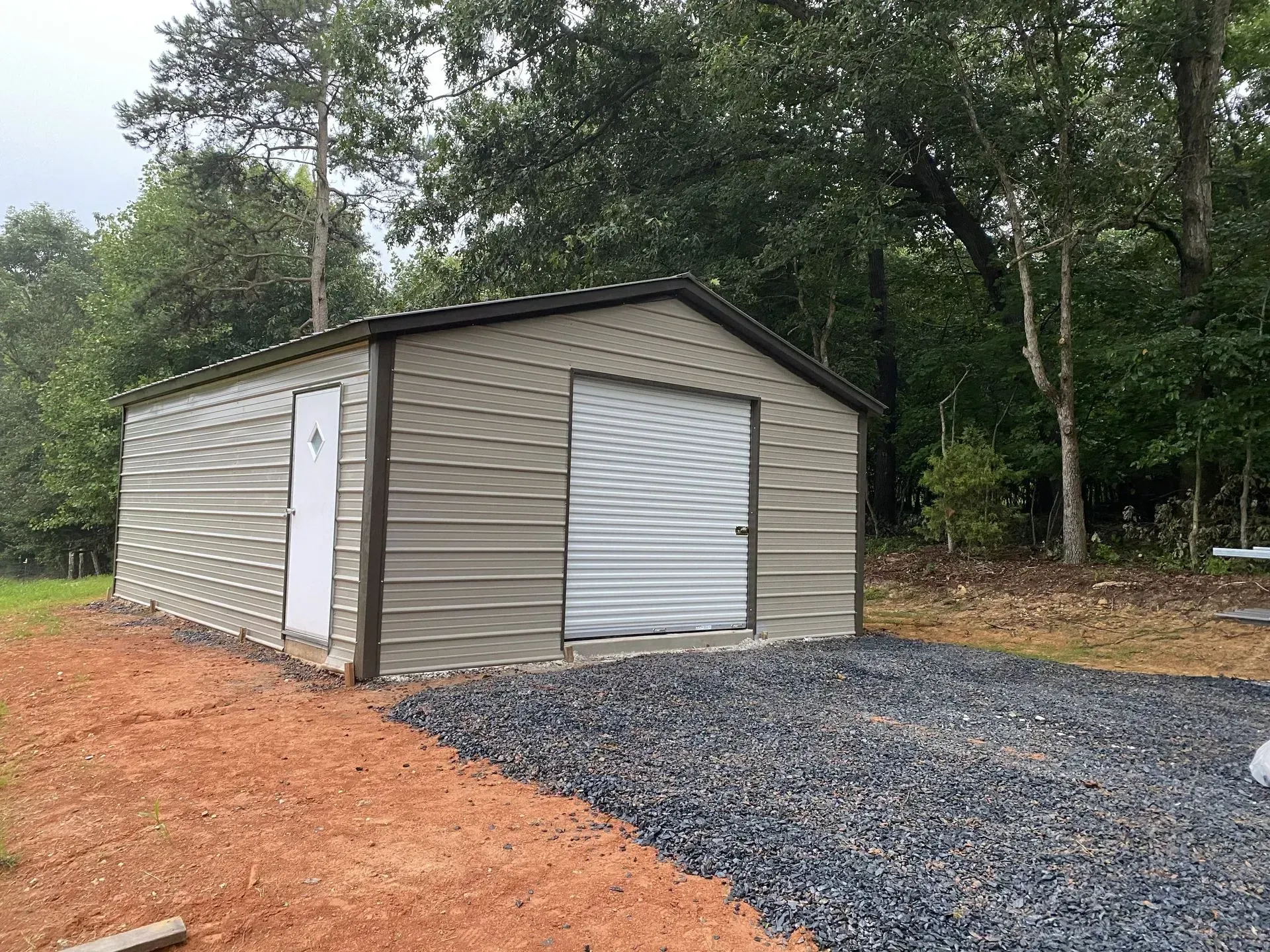 A metal garage with a white door is sitting in the middle of a gravel driveway.
