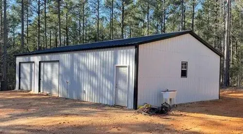 A white metal building with a black roof is sitting in the middle of a dirt field.