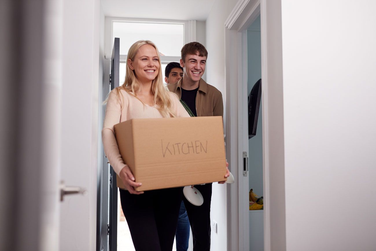 Two people carrying a cardboard box through a hallway, smiling.
