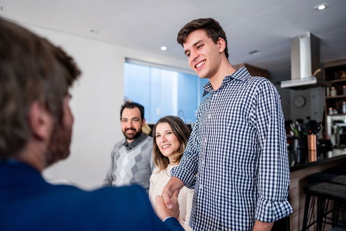 Three people chatting in a bright kitchen, one standing and smiling while shaking hands with someone seated.