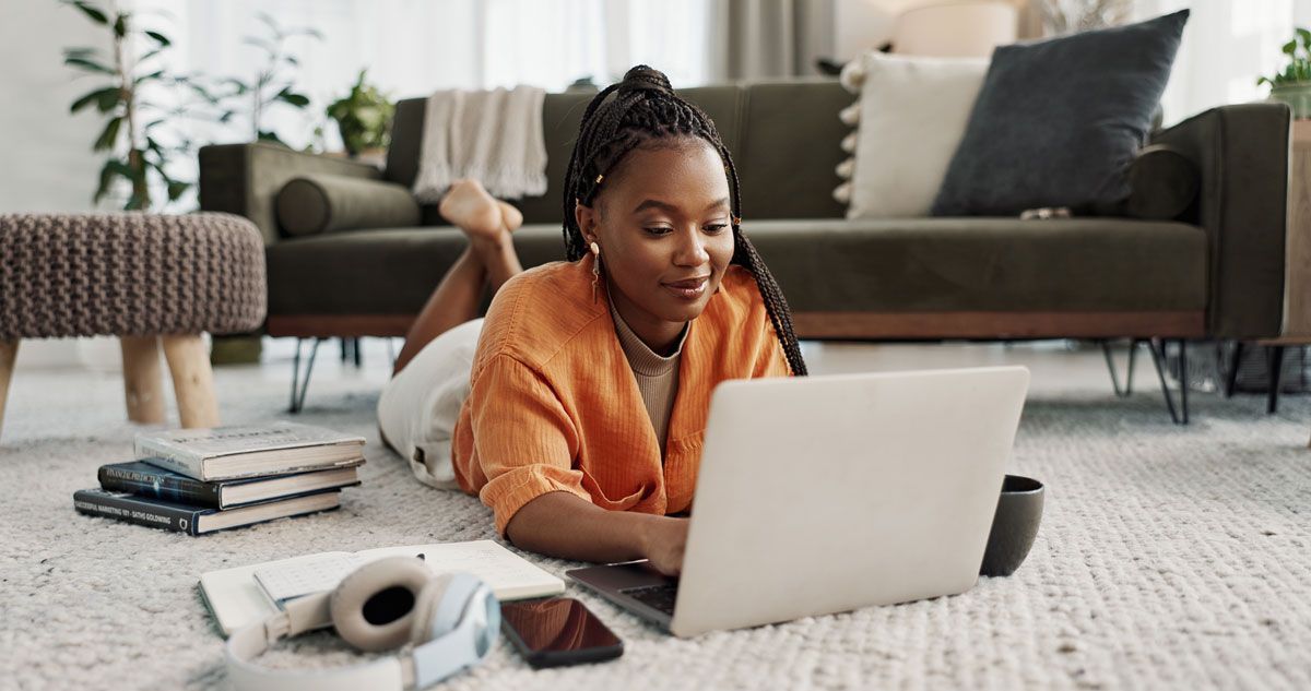 Person lying on a rug using a laptop in a cozy living room with books, a plant, and a sofa.